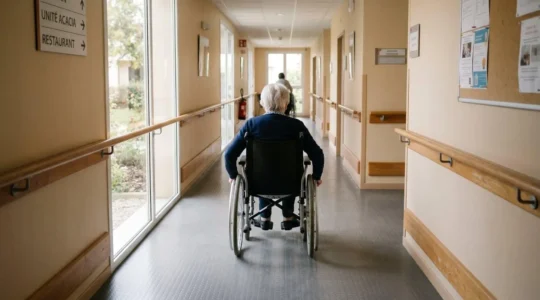 Personne âgée en fauteuil roulant circulant de manière autonome dans un couloir d'EHPAD équipé de mains courantes et d'un sol antidérapant
